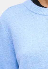 Κοντινό πλάνο ενός ατόμου που φοράει ανοιχτό μπλε πλεκτό πουλόβερ με ριμπ πλεκτό λαιμόκοψη πάνω από μαύρο εσωτερικό ρούχο.