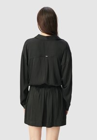 Schwarzes Kleid mit langen Ärmeln, einem Kragen und einer gerafften Taille. Der Stoff wirkt glatt mit leichtem Glanz und zeichnet sich durch ein dezentes Faltendetail auf der Rückseite aus.
