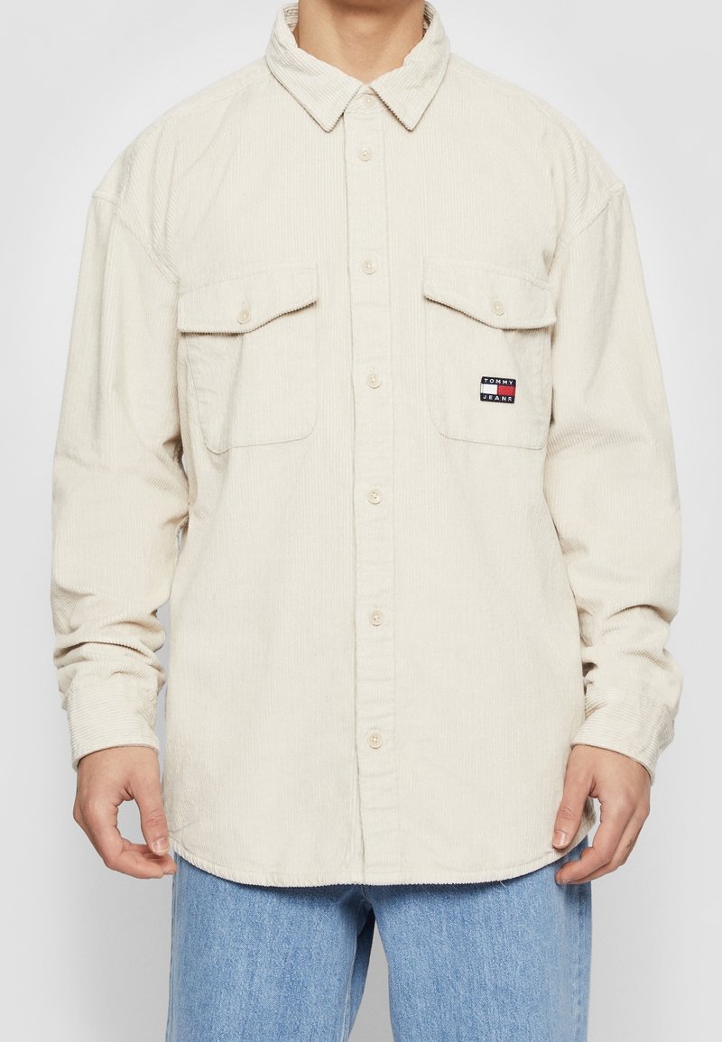 Beiges Cordhemd mit Knöpfen und zwei Brusttaschen, eine davon mit einem Tommy Jeans Logoaufnäher, kombiniert mit blauer Jeans.