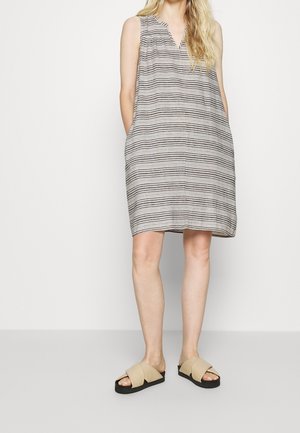 Mujer con vestido sin mangas a rayas hasta la rodilla y sandalias beige, de pie sobre un fondo claro y liso.