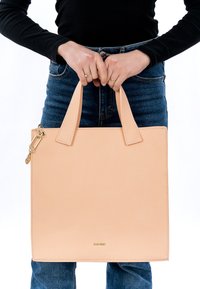 Persona che indossa una maglia scura a maniche lunghe e jeans blu, che tiene una grande borsa rettangolare in pelle beige per i manici.