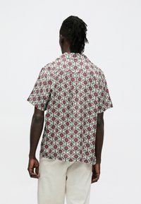 Koszula z krótkim rękawem w kremowej tkaninie z geometrycznym wzorem w czerwono-zielone kolory. Luźny fason z kołnierzem typu camp oraz prostym dołem.