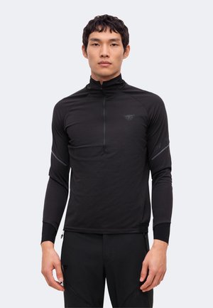 Un homme porte un haut de sport noir à manches longues avec une fermeture éclair courte, ainsi qu’un pantalon noir, sur un fond uni clair.