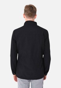 Maglietta nera a maniche lunghe con colletto dritto e vestibilità rilassata, realizzata in tessuto morbido con una leggera lucentezza. Vista posteriore mostrata.