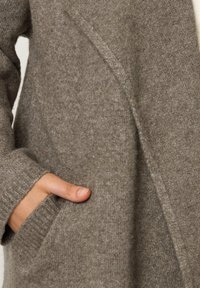 Cardigan grigio in misto lana con una texture morbida, polsini a coste e tasche laterali. Il design aperto mette in risalto linee pulite e dettagli minimi.