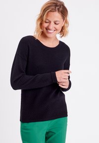 Pull noir texturé avec un col rond et des manches longues, présentant un motif en nid d'abeille. Assorti à un pantalon vert.
