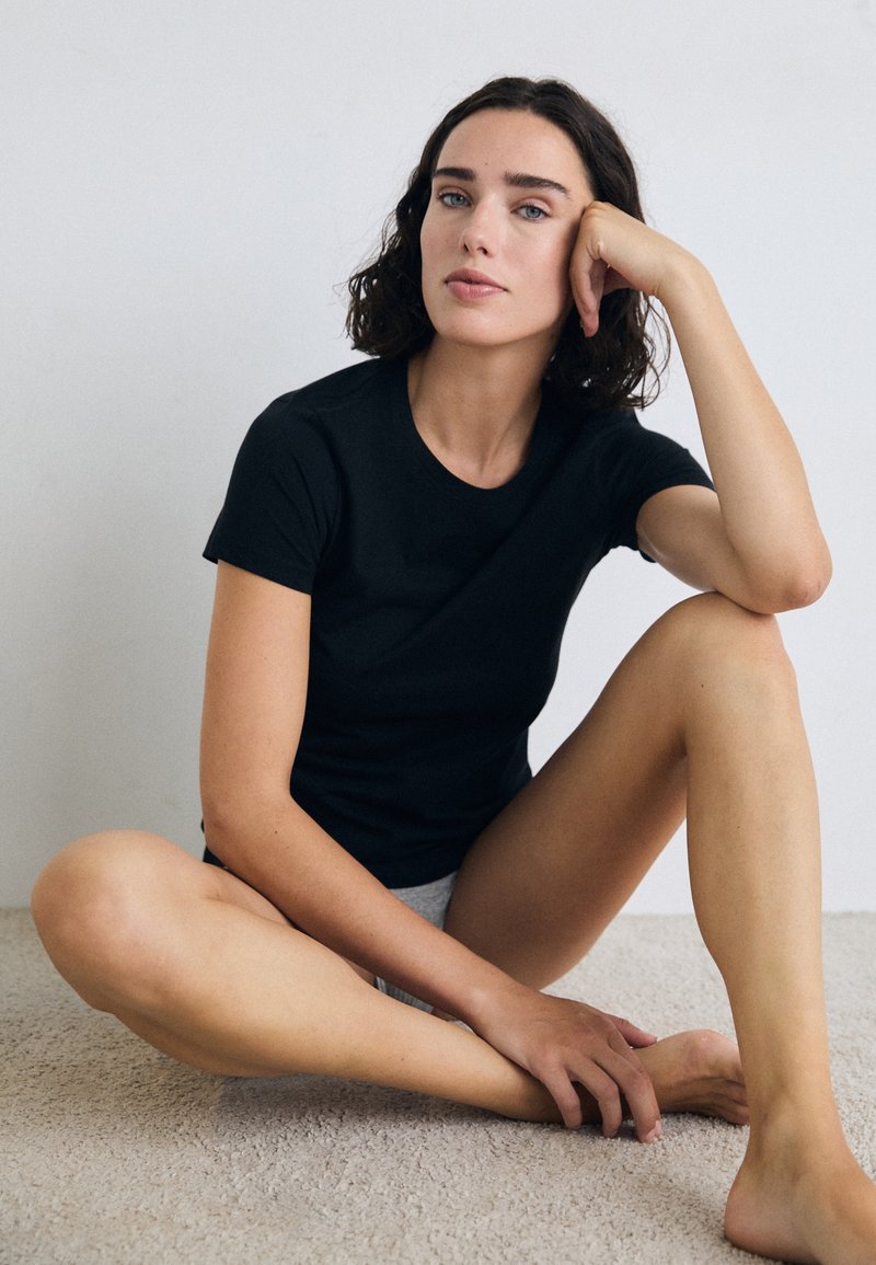 Une femme aux cheveux bouclés mi-longs est assise sur un tapis, portant un t-shirt noir et un short gris, reposant son menton sur sa main.