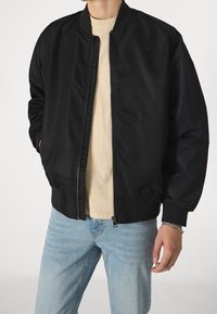 Chaqueta bomber negra con cierre delantero, cuello y puños acanalados, tela suave y corte relajado. Usada sobre una camisa beige clara con jeans azules.