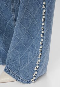 Džínová nohavica s diamantovým prešívaním a dekoratívnym vertikálnym radom strieborných a perlových korálok pozdĺž vonkajšieho švu, nosená s bielou špicatou obuvou.