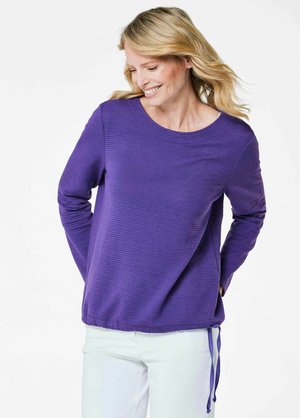 Lila Langarmshirt mit gerippter Textur, runder Halslinie und verstellbaren Bindebändern am Saum; getragen mit hellfarbigen Hosen.