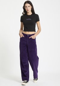 T-shirt noir court avec texte brodé violet ; pantalon en velours côtelé violet à jambe large ; modèle avec les mains dans les poches, portant des baskets noires.