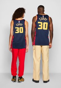 Maglie da basket blu marina con accenti rossi, con "CURRY" e "30" in giallo sul retro. Si indossano pantaloni della tuta rossi e pantaloni cargo beige.