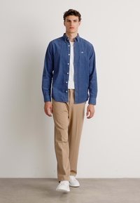 Blauwe button-up shirt van zachte stof, gecombineerd met beige broeken en witte sneakers; model staat voor een neutrale achtergrond.