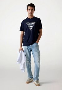 T-shirt di cotone blu navy con logo triangolare argento e scritta, abbinata a jeans azzurri e sneaker beige. Tiene in mano una felpa bianca.