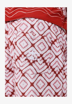 Camiseta roja con líneas blancas combinada con pantalones cortos blancos que presentan un patrón de rombos rojos. Un logo blanco bordado se encuentra en el bolsillo trasero de los pantalones cortos.