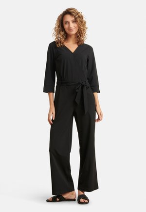 Zwart wikkel jumpsuit met driekwart mouwen, strikdetail bij de taille en brede pijpen; gedragen met platte zwarte sandalen.