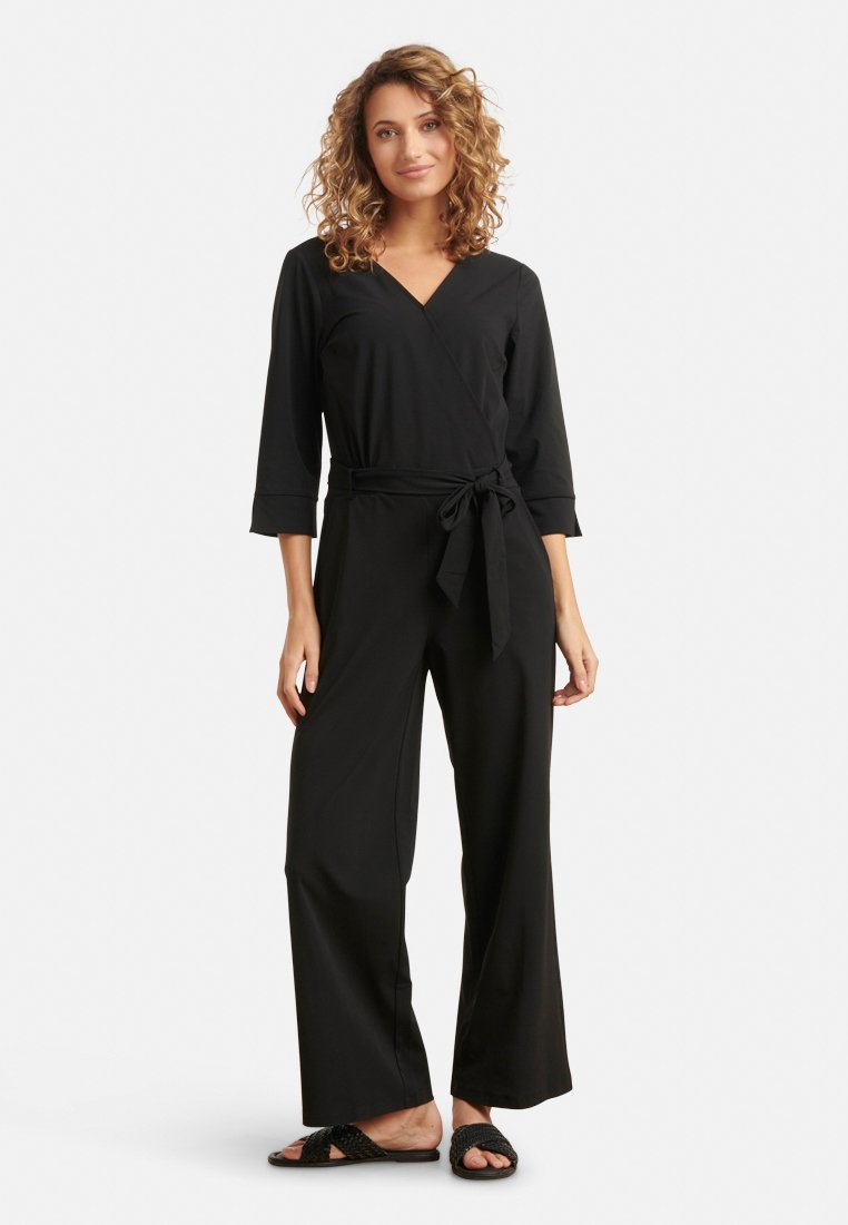 Schwarzer Maxi-Overall mit Dreiviertelärmeln, Taillenknoten und weitem Beinschnitt; getragen mit flachen schwarzen Sandalen.