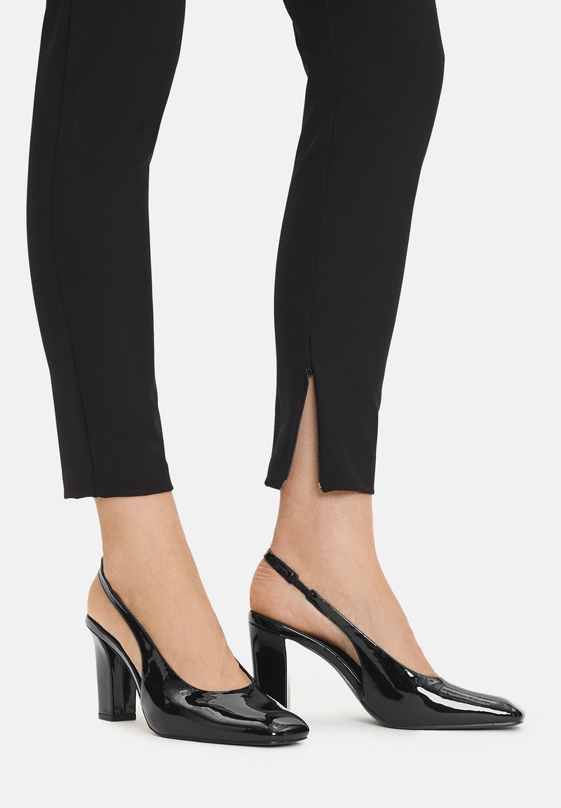 Czarne lakierowane buty na obcasie typu slingback z ostrym noskiem, masywnym obcasem i wyraźnym łukiem. Noszone z dopasowanymi czarnymi spodniami z detalem zamka.