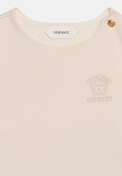 Κρέμ τοπ Versace με στρογγυλή λαιμόκοψη, χρυσό λογότυπο Medusa από μικρές χάντρες και ένα χρυσό διακοσμητικό κουμπί στον ώμο.
