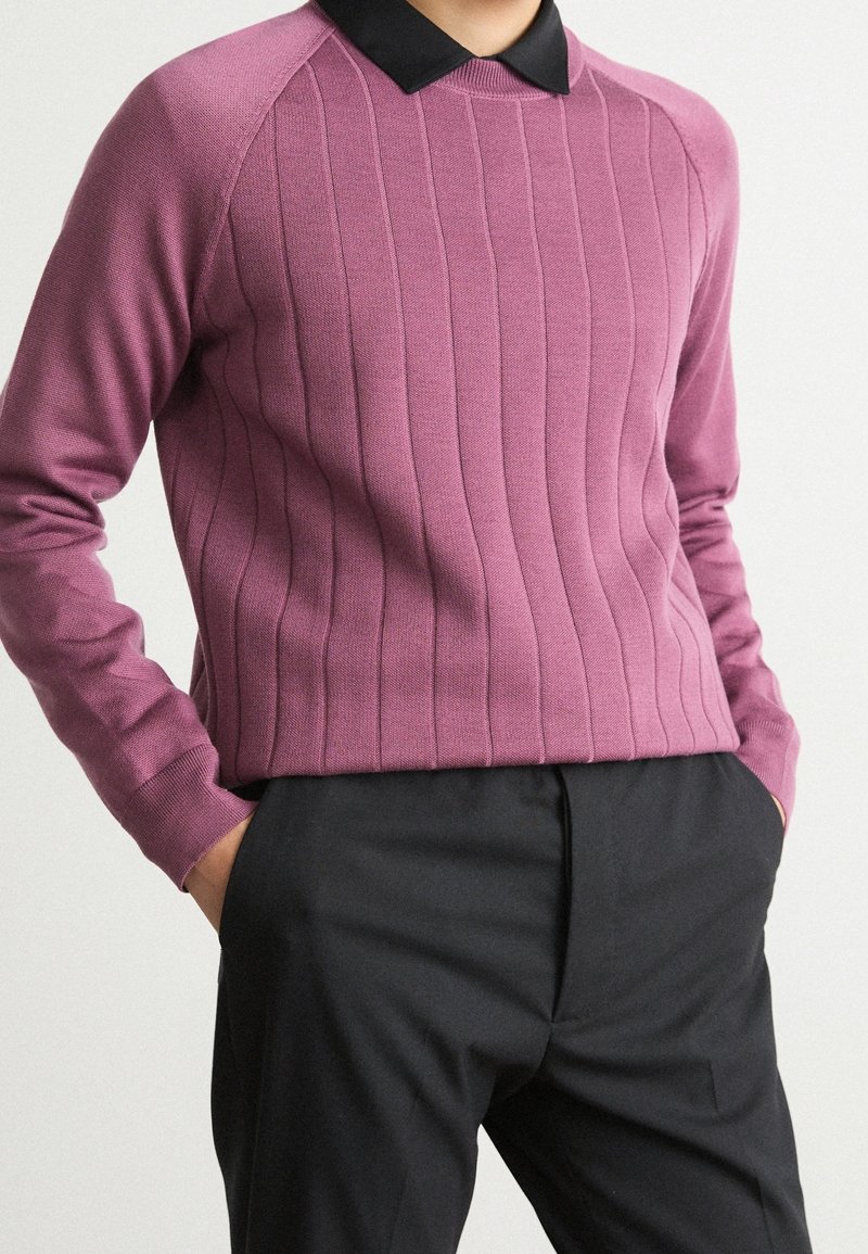 Personne portant un pull côtelé mauve sur une chemise noire à col, les mains dans les poches d'un pantalon noir, sur un fond uni.