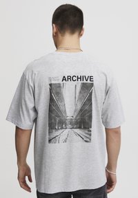 Grå bomull t-skjorte med et stort svart-hvitt fotografisk trykk av en arkitektonisk scene på baksiden, med "ARCHIVE" tekst over.