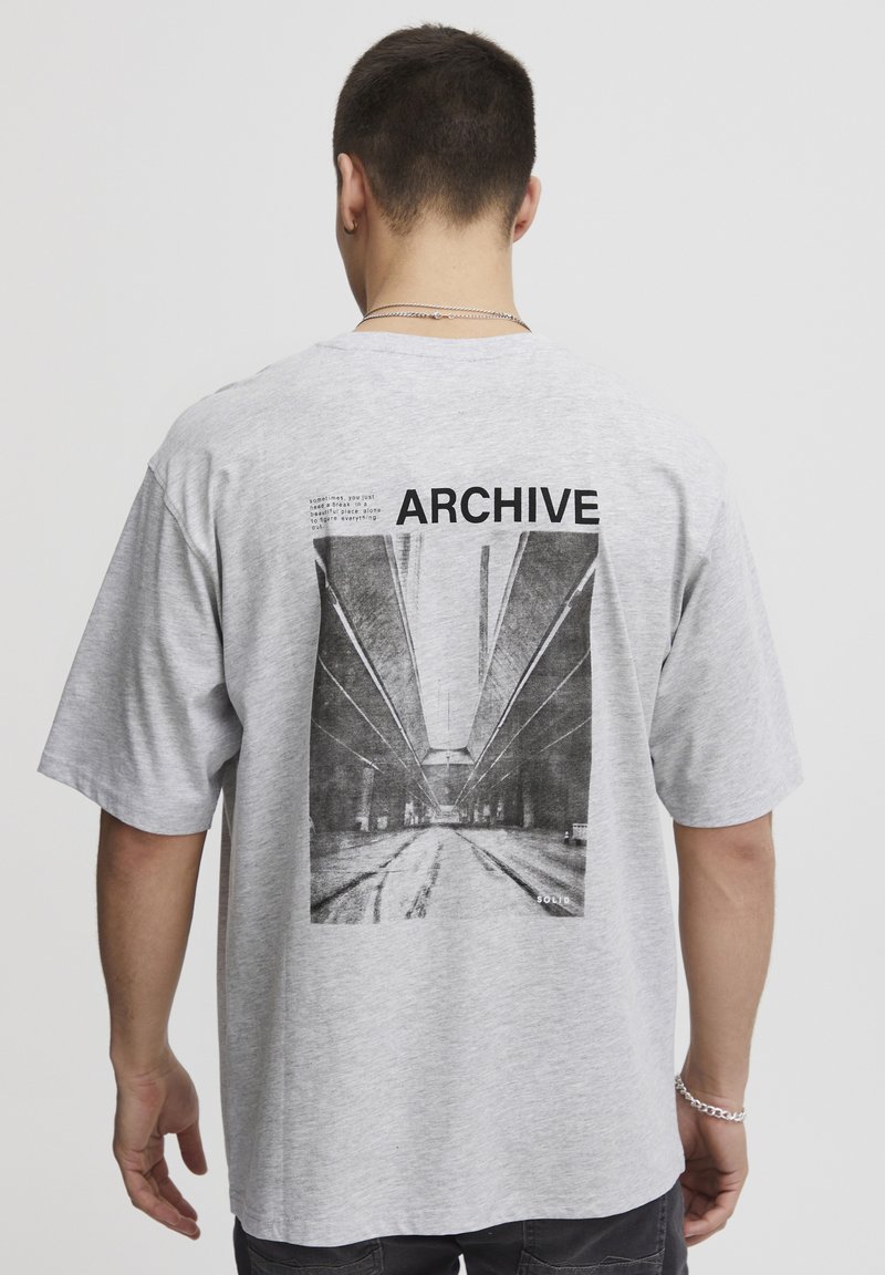 Grå bomull t-skjorte med et stort svart-hvitt fotografisk trykk av en arkitektonisk scene på baksiden, med "ARCHIVE" tekst over.