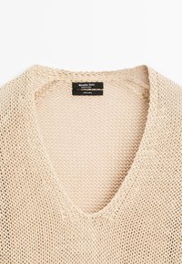 Beżowy sweter z dekoltem w serek wykonany w 100% z lnu, z luźnym splotem i delikatnym prążkowanym wykończeniem przy dekolcie i dolnej krawędzi.