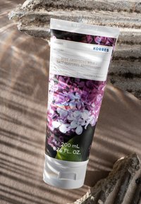 Vit tub med en blomsterdesign i lila, märkt "Body Smoothing Milk," med en svart bakgrund och ett vitt skruvkork. 200 mL.