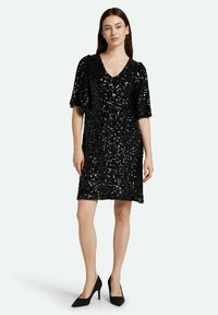 Vestido negro de lentejuelas con escote en V y mangas cortas, que presenta un corte holgado y una textura brillante. Combinado con zapatos de tacón alto negros.