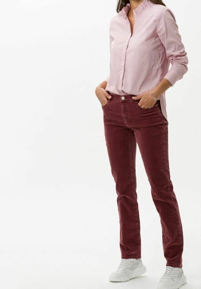 Femme portant une chemise rose clair boutonnée, un pantalon rouge foncé et des baskets blanches, debout les mains dans les poches devant un fond uni.