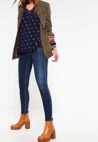 Botas marrones de tacón, vaqueros oscuros ajustados y una blusa azul marino con un patrón rojo disperso, acompañada de una chaqueta verde oliva.