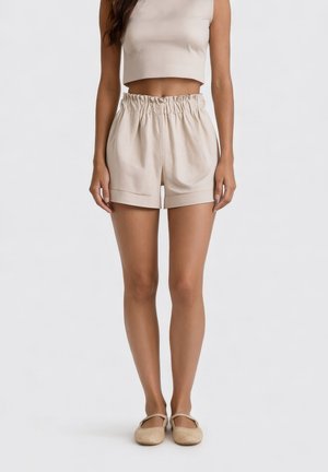 Donna che indossa pantaloncini paperbag beige a vita alta, top corto abbinato e ballerine Mary Jane beige, in piedi davanti a uno sfondo semplice.