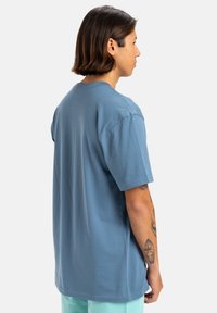 Camiseta de algodón azul con mangas cortas, cuello redondo y corte relajado. La vista trasera muestra una textura suave y costuras planas. Diseño liso sin patrones.