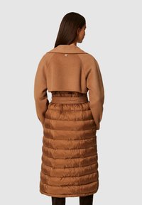 Hnedý skrátený sveter so širokým golierom, cez dlhý, objemný, prešívaný hnedý kabát s opaskom v pase.