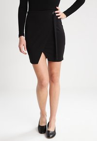 Černá mini zavinovací sukně z pružného materiálu, s bočním vázáním a diagonálním lemem. V kombinaci s černými lodičkami se zavřenou špičkou.