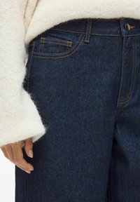 Dark blue denim jeans feature contrasting orange stitching, a front pocket, and a button closure, paired with a textured white sweater sleeve.