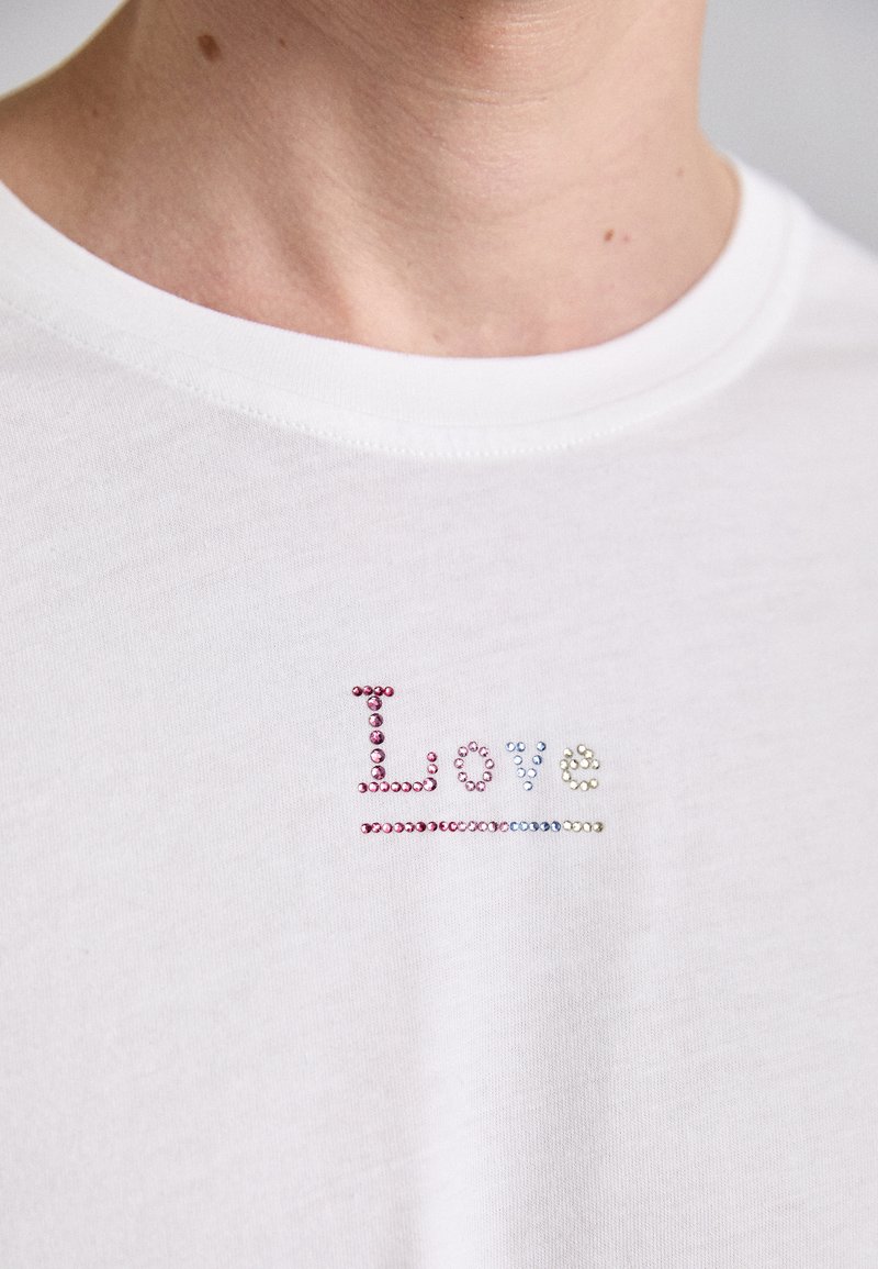 White short-sleeve cotton t-shirt featuring the word “Love” embellished with colorful rhinestones in pink, blue, and clear finishes.
