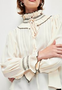 Cremfarbene Bluse mit plissiertem Design, Rüschkragen und schwarzem Saum. Verfügt über lange Puffärmel und Knopfakzente an den Bündchen.