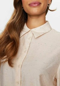 Gai krēms pogu krekls ar mīkstu tekstūru, ar daudzkrāsainiem plankumiem un klasisku apkakli. Redzami septiņi pogas gar priekšpusi.