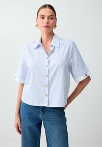 Femme portant une chemise à manches courtes rayée bleu clair et blanc avec des boutons dorés et un jean bleu, debout la main sur la hanche.