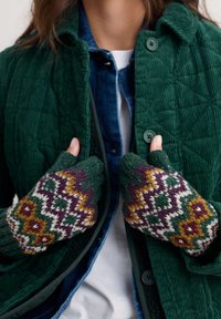 Persona que lleva mitones de lana con un diseño estampado abrochándose los botones de una chaqueta acolchada verde sobre una camisa de mezclilla y una camiseta blanca.