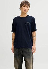 Camiseta de algodón azul marino de manga corta con un logo en el lado izquierdo del pecho. Combinada con unos vaqueros negros desgastados, mostrando un ajuste relajado.