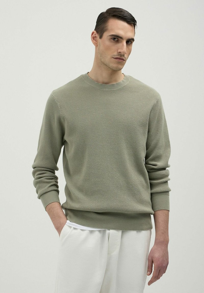 Pull en tricot vert olive clair avec un col rond et des poignets côtelés. Associé à un pantalon blanc, au fit décontracté.