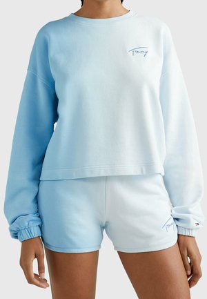 Personne portant un sweat-shirt manches longues dégradé bleu clair et blanc ainsi qu'un short assorti avec de petits logos brodés.