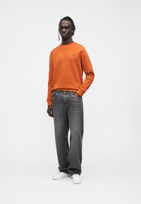 Orange sweatshirt with round neck, small logo on chest, paired with dark gray wide-leg jeans and white sneakers. Smooth texture, casual fit.
