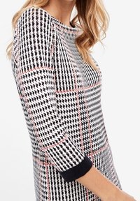 Robe à carreaux noir et blanc avec des accents roses, manches trois-quarts et poignets bleu marine. Texture en tricot et décolleté rond.