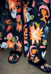 Kukkapainatuksilla varustetut housut tummassa kankaassa esittelevät oransseja, vaaleanpunaisia ja vihreitä kuvioita. Mustissa tossuissa on pehmeä, karvainen reunus ja värikkäitä yksityiskohtia.