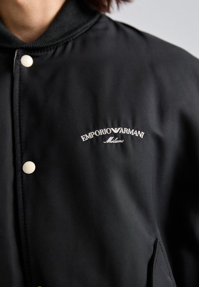 Melna neilona jaka ar ribotu apkakli, spiedpogu aizdari un izšūtu "Emporio Armani Milano" logo kreisajā krūtīs.