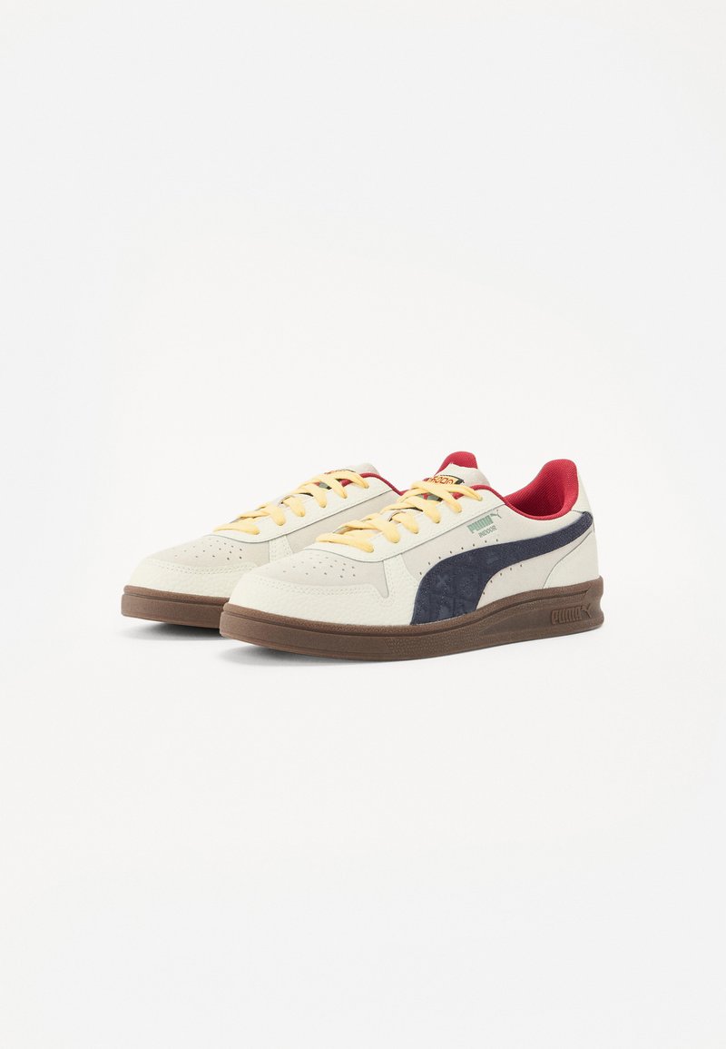 Jasne kremowe sneakersy z granatowymi akcentami, żółtymi sznurówkami i czerwonym wnętrzem. Posiadają perforowane detale oraz brązową gumową podeszwę.