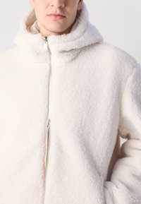 Krem boje flis jakna s kapuljačom, koja ima vidljivi patentni zatvarač i teksturiranu površinu. Jednostavan dizajn koji pruža toplinu i udobnost.